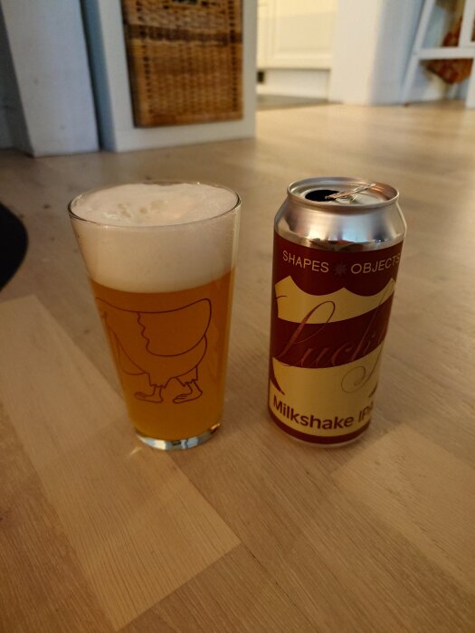 Ett glas öl med skum bredvid en öppen burk märkt "Shapes Objects Lucky Star Milkshake IPA" på ett trägolv inomhus.