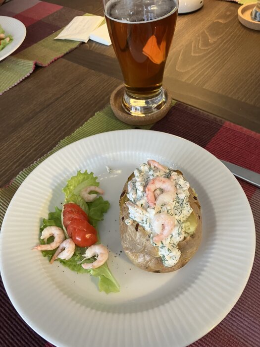 Bakad potatis med skagenröra och räkor, serverad med sallad och tomat på en tallrik, bredvid ett glas med dryck.