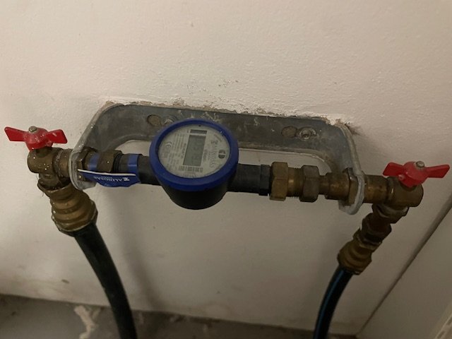 Vattenmätare monterad på ett rörsystem med två röda ventilhandtag, installerad på en vägg.