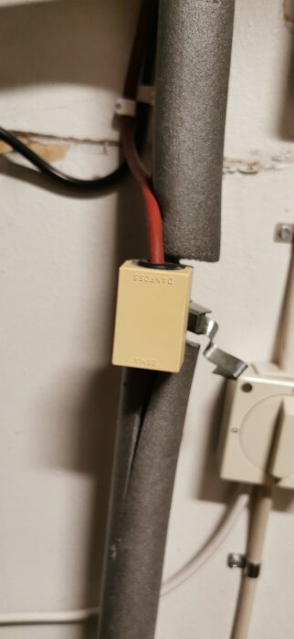 Värmesensor från Danfoss monterad på isolerat rör, med röd kabel och metallklämma, jämte termostat på väggen.