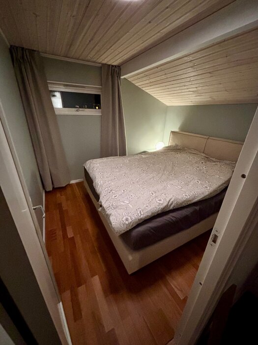 Litet sovrum med snedtak, dubbelsäng med mönstrat överkast, beige väggar, fönster med grå gardiner och mörkt trägolv.