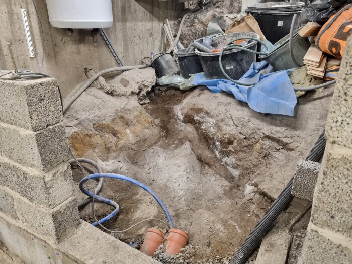 Renoveringsplats med jordutgrävning, markrör och byggmaterial. Cementblock omger rör och verktyg belamrar området.