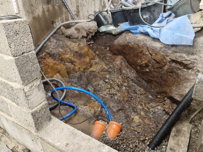 Grävd grop med synliga rör och stenar, omgiven av cementblock och diverse byggmaterial.