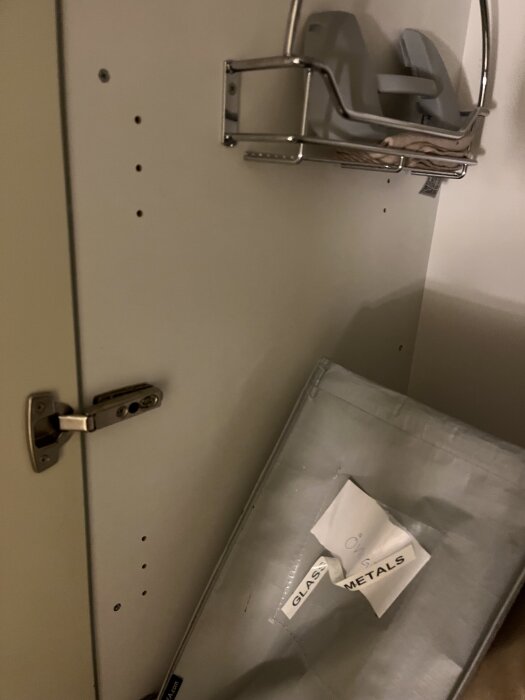 Inuti ett skåp med hål för hyllplacering, en metallkorg och en kasse markerad för glas och metallåtervinning.