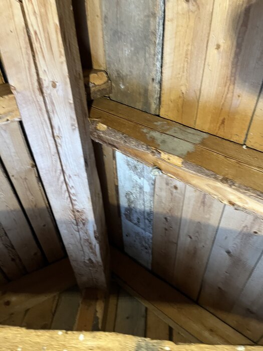 Trästomme och bjälklag på vinden i ett hus från 1930-talet med synliga brädor, färgungar och märken som kan tyda på fukt eller slitage.