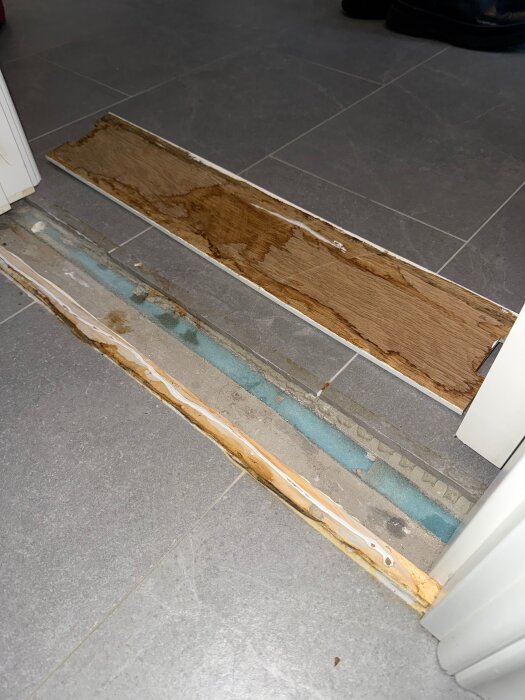 Tröskel vid badrumsdörren med trä och plastmaterial, visar missfärgningar som tyder på fuktproblem. Grått klinkergolv runtom.