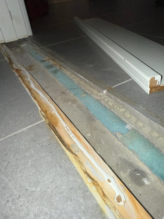 Tröskel på badrumsgolv med missfärgningar och synligt tätskikt. Avtagen tröskel visar blått isoleringsmaterial och bruna fläckar.