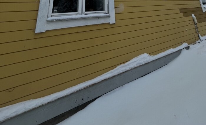 Gul träfasad med vitt fönster och snö vid marken. Möjlig rötskada på syll under fönstret.