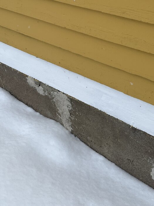 Gul träfasad med vitmålad syll vid snötäckt mark, tecken på slitage synliga på betongen.