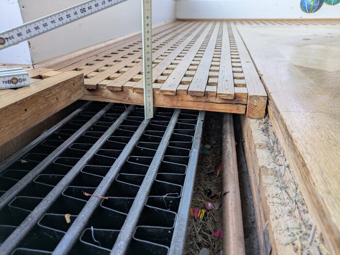 Måttstock mäter avstånd från träpanel till underliggande galler och rör, visar begränsat utrymme för fläktinstallation.