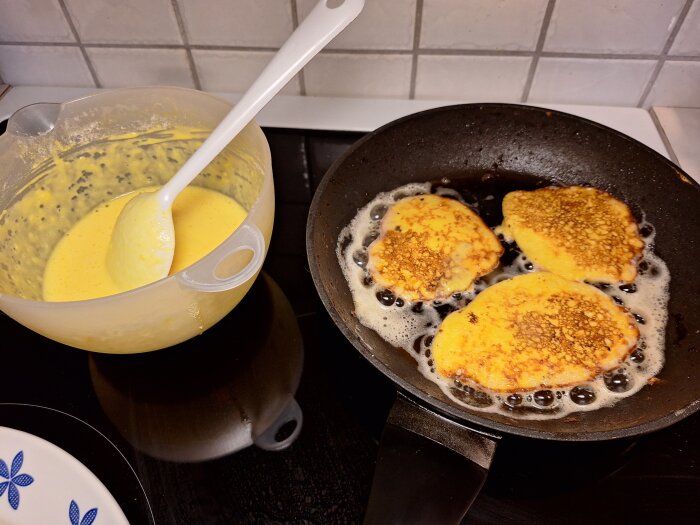 Pannkakor steks i en stekpanna bredvid en skål med pannkakssmet och en slev på en spis.