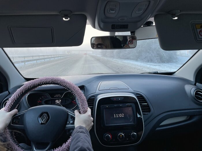 Bilinteriör, en person håller i ratten med ett rosa överdrag. Vägen utanför är snötäckt och disig med en hastighet på 92 km/h. Klockan visar 11:02.