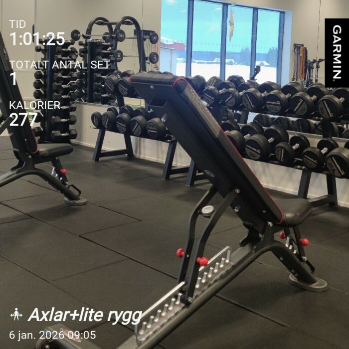 Träningsbänk på gym med hantlar och spegelvägg i bakgrunden, del av träningspass visas på display från Garmin-enhet.
