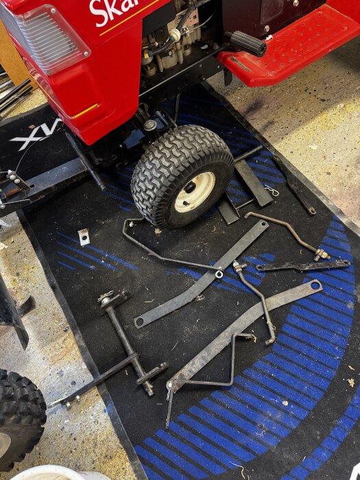 Delar från en Skandi trädgårdstraktor med snöblad ligger utspridda på marken bredvid traktorn i ett garage, inklusive hjul och metallstänger.