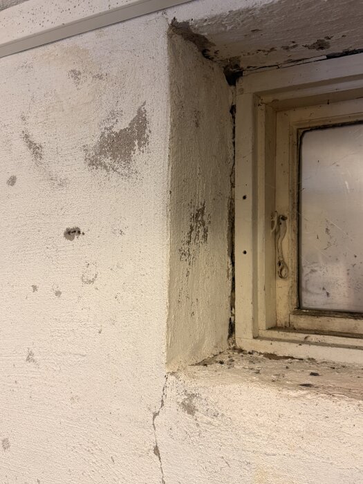 Vägg med flagnande puts och sprickor runt ett källarfönster, behov av renovering och målning.