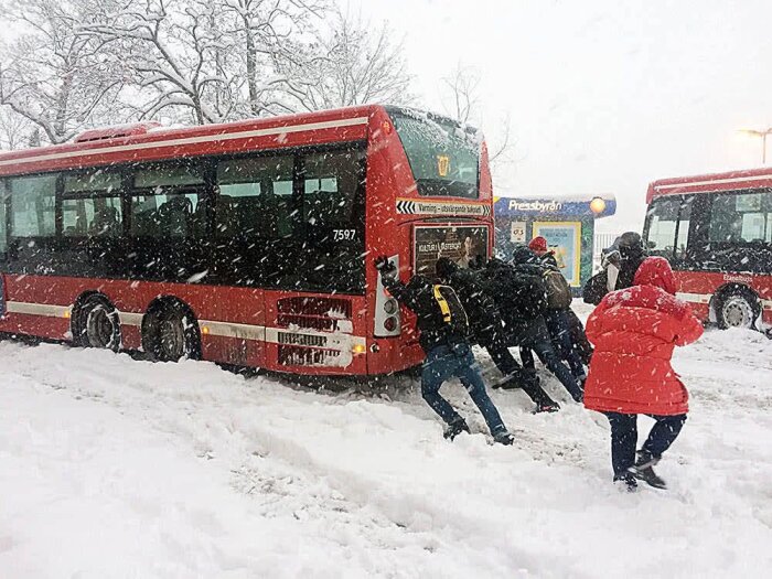Människor hjälper till att skjuta en röd buss genom snöfall på en snöig väg.