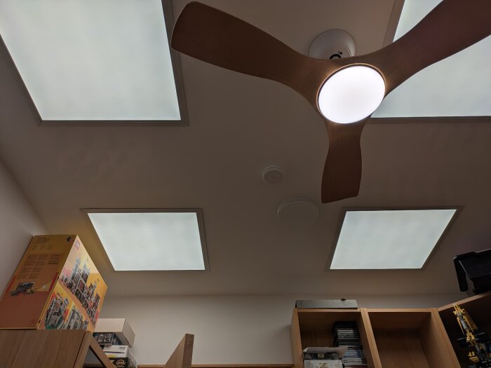 LED-paneler i taket med varm gul belysning i ett hemmakontor, synliga hyllor och takfläkt.