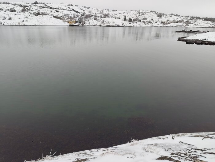 Klart vatten i vinterlandskap med snötäckta kullar i bakgrunden och en liten gul byggnad vid strandkanten.