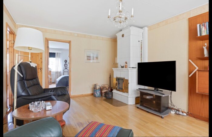 Vardagsrum med svart TV, vit öppen spis, läsfåtölj och kristallkrona. Trägolv och dörrar, moderna och retro detaljer, mysig atmosfär.