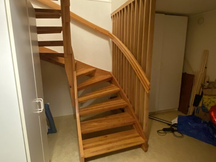Trätrappa i ett rum med vita väggar och en garderob, med öppna hyllsteg och handledare. Golvet är beige och det finns byggmaterial vid sidan.