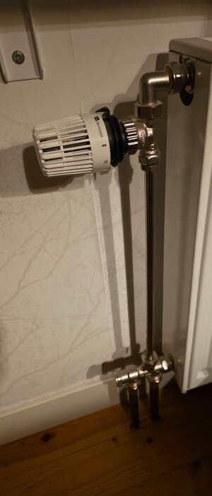 Radiator med termostatventil installerad mot en ljus vägg, kopplad med metallrör ner till golvet av trä.