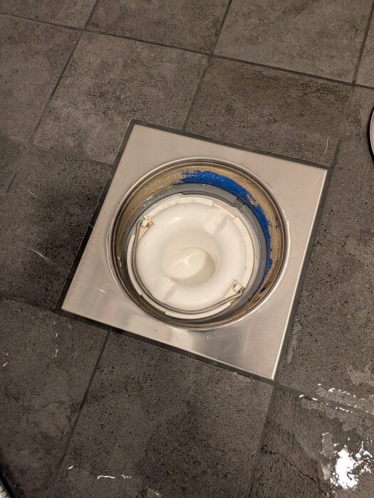 Vattenlås i duschgolv med delvis blå beläggning runtom på grått kakelgolv.