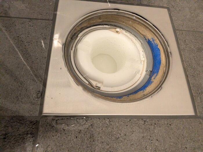 Vattenlås i duschgolv med synlig blå tätning och beige smuts runt kanterna, monterat på en grå kaklad yta.