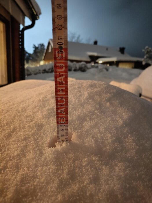 Måttstock visar snödjup på cirka 50 cm i en trädgård under kvällen. Snö har täckt tak och omgivande landskap.