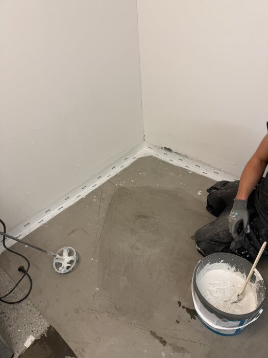 Arbetare på knä vid golv i pannrum. Vägghörn med tätskikt och murbruk i en hink. Verktyg och material förberedda för fogning.
