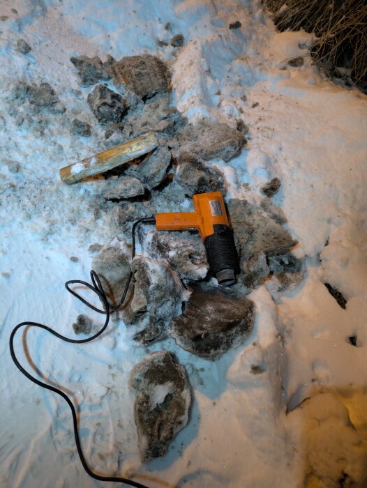 En orange varmluftspistol ligger på snöig mark, omgiven av stenar och en träbit.