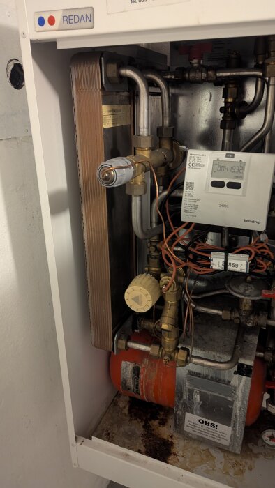 Uppvärmningssystem med rörledningar, ventiler och mätare i en vit metallbox, med märkning och kablage synligt.