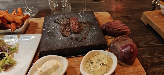 Grillad köttbit på het stenplatta med två såser och sötpotatispommes på ett bord i Borlänge.