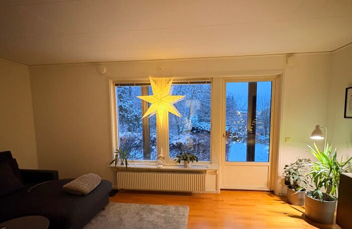 Vardagsrum med stort fönster, julstjärna hängande, utsikt mot snötäckt trädgård och soffhörna med växtarrangemang.