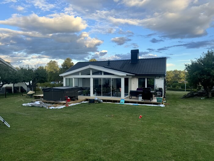 En villa med ett stort uterum av glas på en grön gräsmatta, med en altan och träd i bakgrunden under en blå himmel med moln.