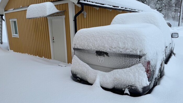 Snötäckt bil parkerad bredvid ett gult hus, ytor runt omkring är täckta av tung snö efter nyligen plogad gårdsplan.