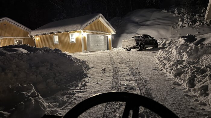 Snötäckt uppfart framför garage och bil parkerad i vinterlandskap, omgiven av snöhögar efter plogning, kvällstid.