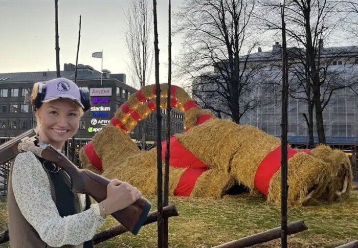 Kvinna med gevär framför halmbock dekorerad med röda band, omgivning av stad och byggnader i bakgrunden.