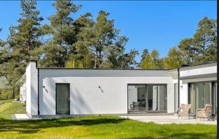 Modernt, vitt attefallshus med platt tak, stora fönster och altandörr, omgiven av träd och grön gräsmatta i soligt väder.