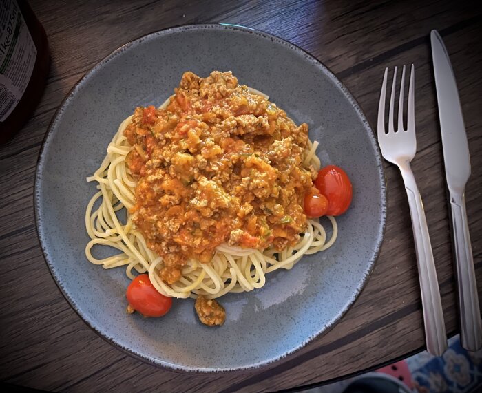 Tallrik med spaghetti toppad med köttfärssås och småtomater på ett träbord, bredvid gaffel och kniv.
