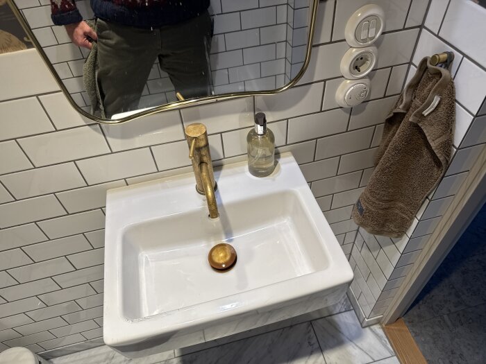 Handfatet Duravit Vero med mässingskran, spegel ovanför, handduk och flaska med flytande tvål på vitkaklad vägg.