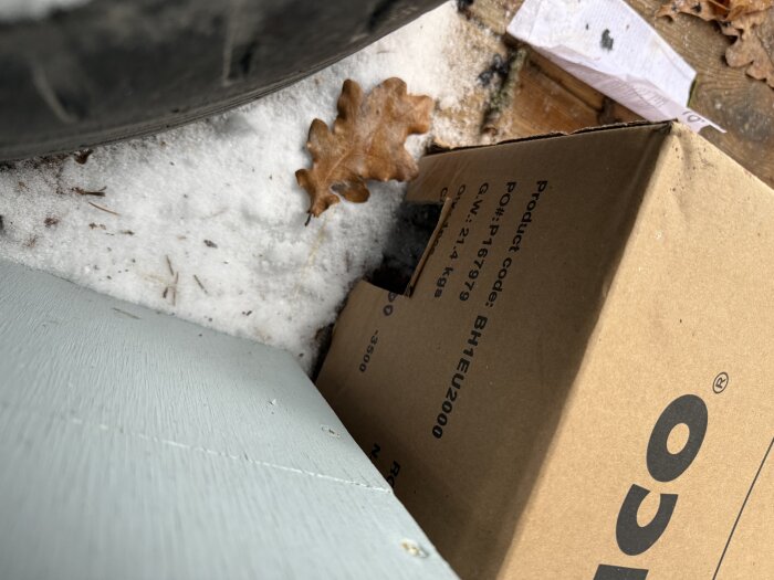 Kartong med tryckt text står på snöig mark bredvid några bruna höstlöv och ett blågrönt träföremål.