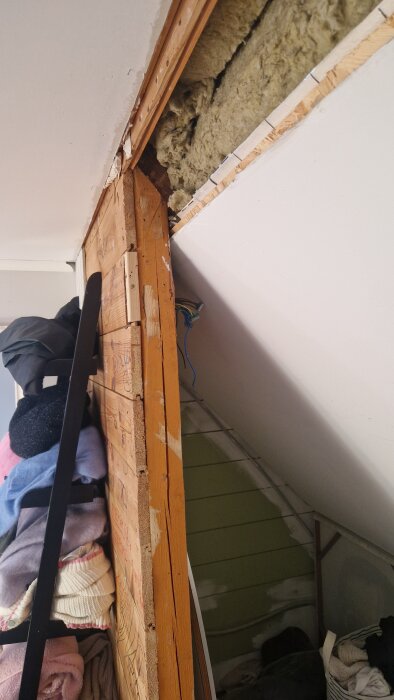 Vägg med synliga träreglar och upphängda kläder, rymmer isolering ovanför.