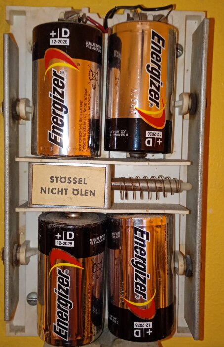 Fyra Energizer D-batterier monterade i en batterihållare med texten "STÖSSEL NICHT ÖLEN" i mitten, mot en gul vägg.