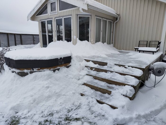 Inglasad altan och badtunna täckt av snö. Snöig terrass med trappa leder till inglasningen.