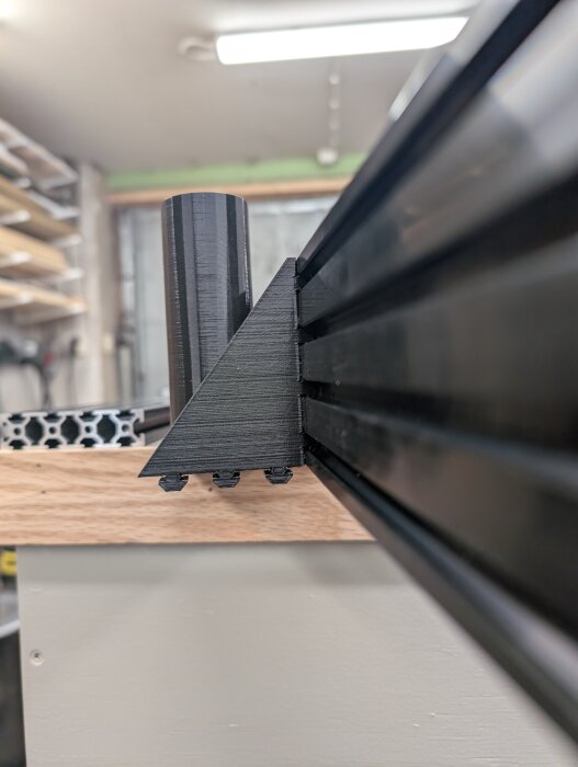 3D-utskriven hållare monterad på en träyta, med en metallprofil i en verkstadsmiljö.