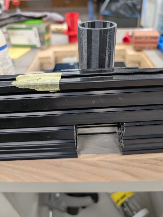 Svart aluminiumbalk med plastcylinder ovanpå, placerad på en träyta, med tejp och verkstadsmaterial i bakgrunden.