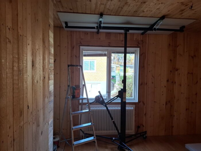 Rum med träpanel, stege och skivlyft för gips i taket. Fönster med utsikt utomhus.