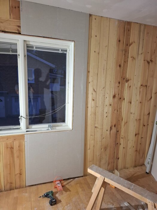 Renovering av rum med gipsskivor på väggarna, fönster med utsikt över hus utanför, verktyg på golvet och arbetsbänk i förgrunden.