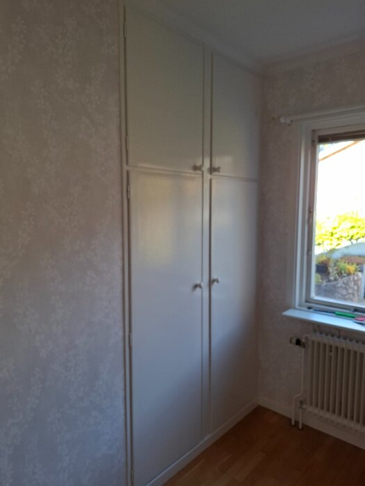 Vit garderob med skjutdörrar, mot en tapetserad vägg i dotterns rum. Detaljer runt fönstret och elementet syns också.