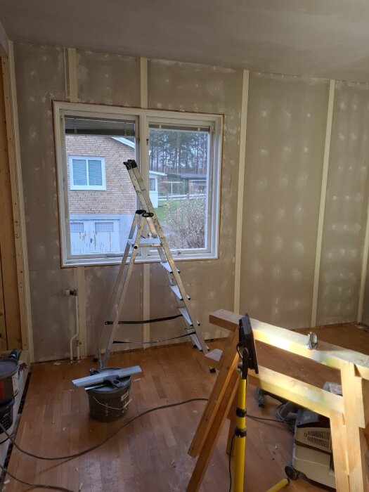 Ett rum under renovering med gipsskivor på väggarna, en stege framför ett fönster och verktyg på golvet.